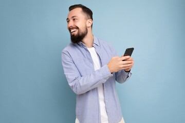Portrait of smiling handsome young brunette unshaven man with beard wearing stylish white t-shirt and blue shirt isolated over blue background with empty space holding in hand and using phone
