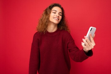 Attractive charming young smiling happy woman holding and using mobile phone taking selfie wearing stylish clothes isolated over wall background