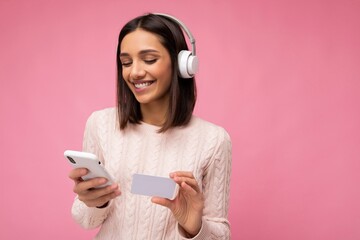 Photo of beautiful young brunette woman wearing pink casual sweater isolated over pink background wall wearing white bluetooth wireless headphones and listening to music and using mobile phone making