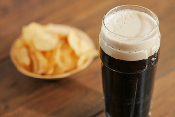 White foam of dark stout beer in focus, portion of potato crisps on a wooden table out of focus....