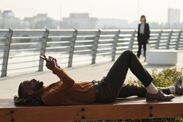 Horizontal side view shot of modern African American man relaxing on wooden bench outdoors watching something on smartphone
