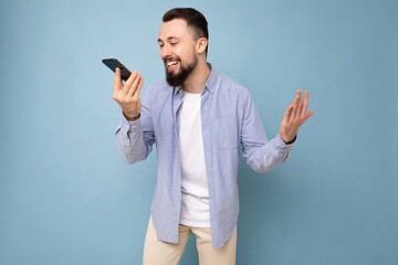 Photo shot of handsome positive good looking young man wearing casual stylish outfit poising isolated on background with empty space holding in hand and using mobile phone recording voice message