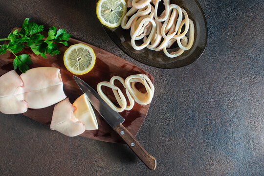 Boiled seafood squid sliced into rings on a wooden board, with lemons and herbs on a brown background.