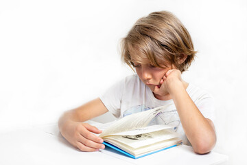 boy reads an interesting book at his desk against the background of a white wall, back to school, education.