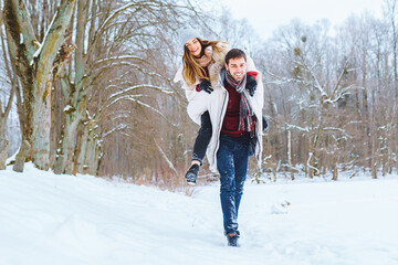 Fototapeta na wymiar Happy young man carry his smiling pretty woman on his back at winter park. Boyfriend giving piggyback ride on shoulder to his girlfriend in winter holiday.