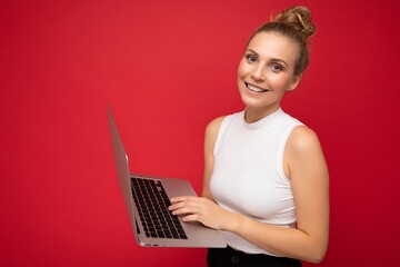 Naklejka premium Side profile photo shot of beautiful smiling blond young woman with gathered hair wearing white t-shirt holding computer laptop typing on keyboard looking at camera isolated over red wall background