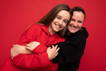 Photo shot of beautiful positive smiling brunette teenager wearing stylish red hoodie and adult brunette woman wearing trendy black hoodie standing isolated over red background wall looking at camera