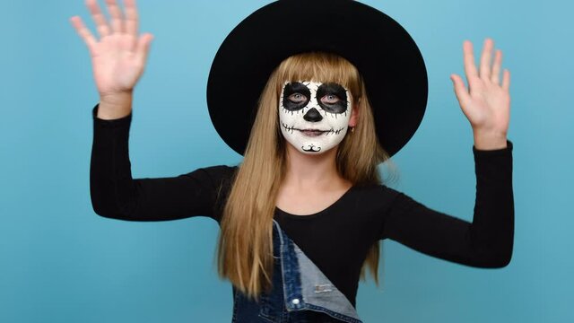 Happy smiling girl kid with Halloween makeup, dance gesticulating hands fool around have fun enjoy, wears big black hat, posing isolated over blue color background in studio. Party holiday concept
