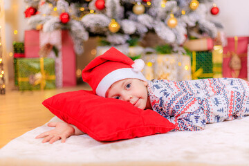 Obraz na płótnie Canvas Boy at the Christmas tree. The boy folded his hands.. Cute baby . Holiday. Article about new year and Christmas. Colorful spruce. Holiday decoration and decor . Santa hat.