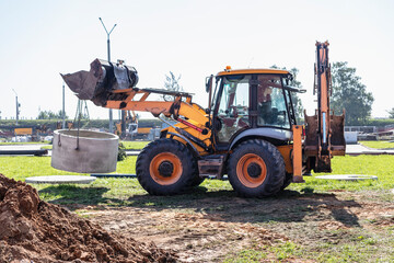 Hydraulic piston system of the excavator with a bucket, lowering into the pit on steel cables concrete sewer ring. Construction or repair of sewer home.