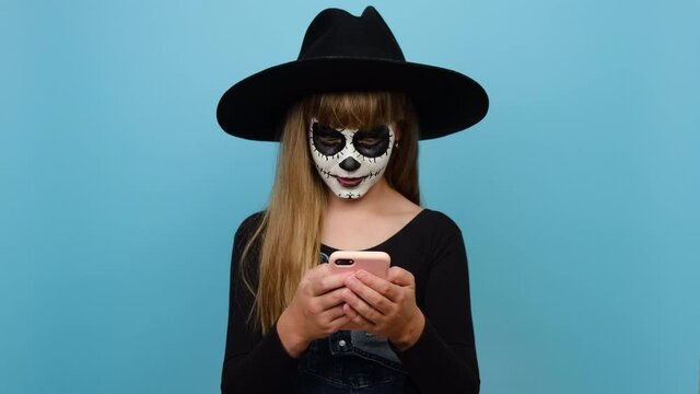 Portrait of satisfied smiling girl child with Halloween makeup mask wear big black hat hold in hand use point on mobile cell phone good news, posing isolated over blue color background in studio