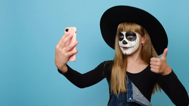 Happy child girl with Halloween makeup mask wears black outfit and hat, get video call using mobile cell phone talk greet with hand, posing isolated on blue color background in studio. Holiday concept