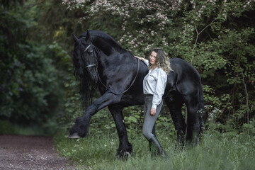 Beautiful young lady posing with a purebred friasian horse in the forest. - 459062595