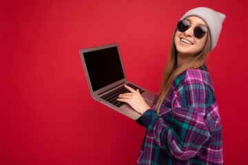 Side profile photo of beautiful smiling happy young woman holding computer wearing casual smart clothes sunglasses and hat looking at camera isolated on red background typing on keyboard text