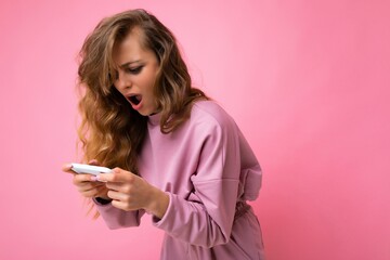 Naklejka premium Side-profile amazed surprised blonde young woman with open mouth wearing pink hoodie isolated over pink background with copy space holding and playing games via telephone looking at device display