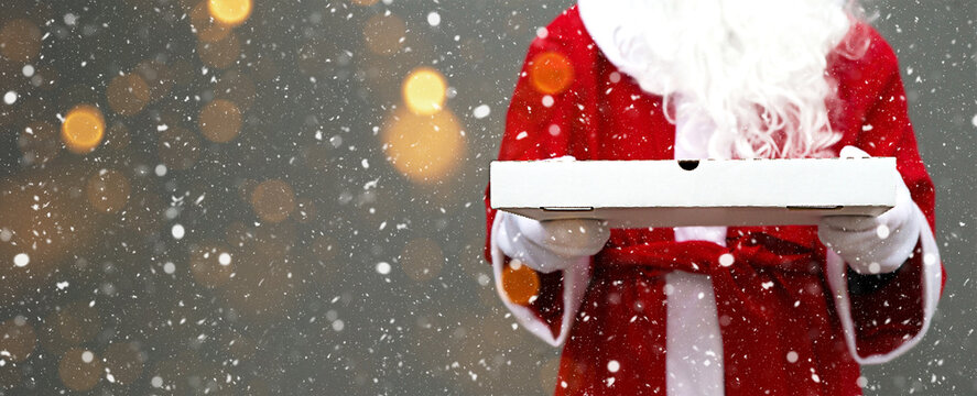 White pizza box in the hands of Santa Claus, with a beard, in a red coat. Christmas fast food delivery. New year's eve promotion. Work on public holidays catering. Copy space, mock up. Banner