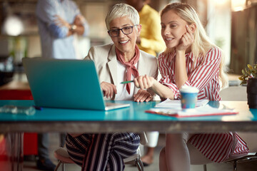 Two female colleagues are sitting at desk in the office watching and commenting laptop content