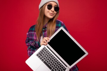 Close-up Photo shot of beautiful smiling dark blond young woman holding computer laptop with empty monitor screen with mock up and copy space wearing sun glasses hat and colourful shirt looking at