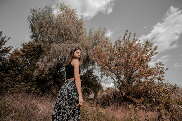 A beautiful slender girl in a long skirt stands against the background of autumn dry trees and a...