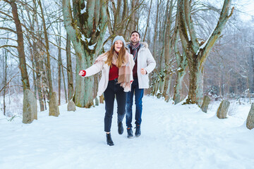 Fototapeta na wymiar Young happy couple smile to each other and walk in winter park holding hands. Man and woman have fun together during winter holidays.