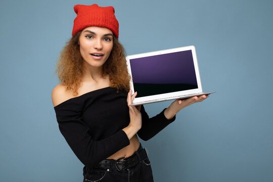 Close-up portrait of surprised funny happy beautiful dark blond woman holding laptop computer looking at camera wearing black crop top and red and orange do-rag isolated over light blue wall