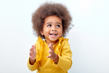Afro fluffy child girl clapping and applauding, happy and joyful, smiling. Adorable charming kid in yellow shirt have fun isolated on white studio background, looking up. Portrait, copy space. - obrazy, fototapety, plakaty
