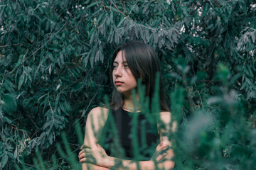 Naklejka premium A young brunette girl has her arms crossed over her chest and looks to the side against a background of dark green leaves. The girl is alone in the forest. High quality photo