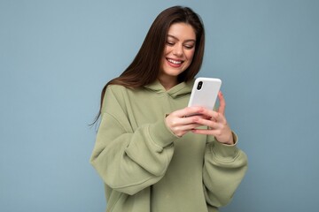 Fototapeta premium Smiling Beautiful young brunette woman wearing stylish green hoodie using mobile phone writing sms isolated on background looking at smartphone screen