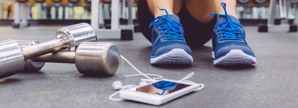 Closeup of unrecognizable sportsman sitting on the floor of the gym with dumbbells and mobile in the foreground