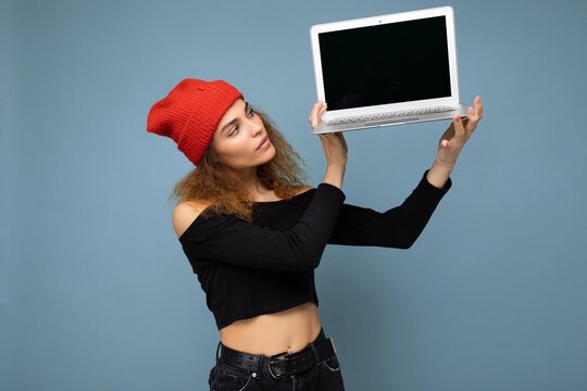 Close-up portrait of beautiful self-confident dark blond curly young woman holding laptop computer looking at netbook keyboard and monitor wearing black crop top and red and orange do-rag isolated