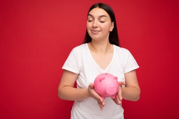 Fototapeta na wymiar Shot of satisfied positive cute young beautiful attractive brunette woman with sincere emotions wearing casual white t-shirt isolated over red wall with empty space and holding pink pig savings box