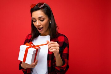 Charming positive surpeised young brunet woman isolated on red background wall wearing white casual t-shirt and stylish red and black shirt holding white gift box with red ribbon and unboxing surprise