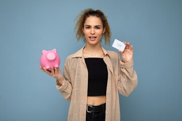 Young dissatisfied asking winsome attractive brunette curly woman with sincere emotions wearing stylish clothes isolated on blue background with copy space, holds pink piggy box and credit card for