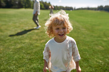 Portrait of adorable little boy smiling away while playing catch together with his parents in green park on a summer day