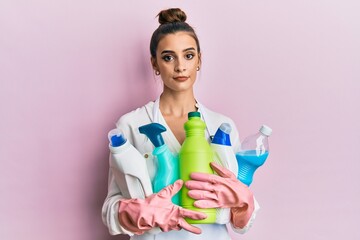 Beautiful brunette young woman wearing cleaner apron holding cleaning products relaxed with serious expression on face. simple and natural looking at the camera.