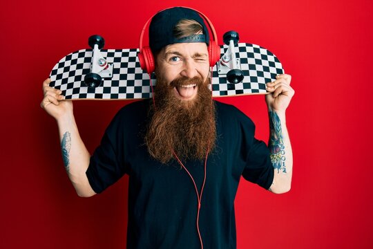 Redhead man with long beard holding skate wearing headphones winking looking at the camera with sexy expression, cheerful and happy face.