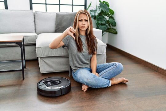 Young caucasian woman sitting at home by vacuum robot with angry face, negative sign showing dislike with thumbs down, rejection concept