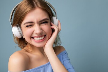 Fototapeta premium Closeup portrait of attractive happy smiling young blonde woman wearing blue crop top isolated on blue background wearing white wireless bluetooth headphones listening to cool music and having fun