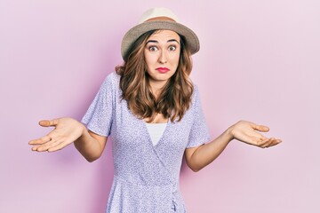 Young hispanic girl wearing summer hat clueless and confused expression with arms and hands raised....