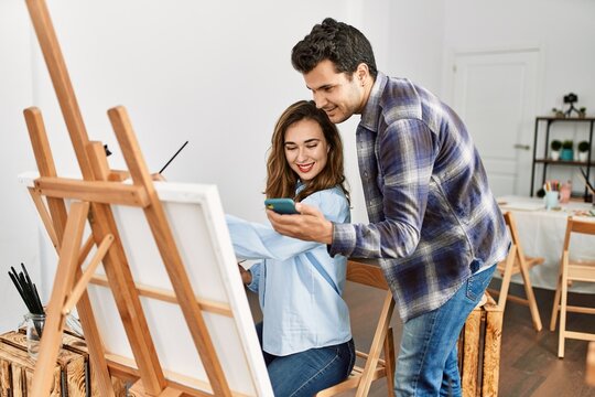 Two hispanic students smiling happy painting and using smartphone at art school.