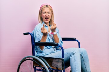 Beautiful blonde woman sitting on wheelchair showing middle finger doing fuck you bad expression,...
