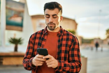 Young hispanic man with serious expression using smartphone at the city.