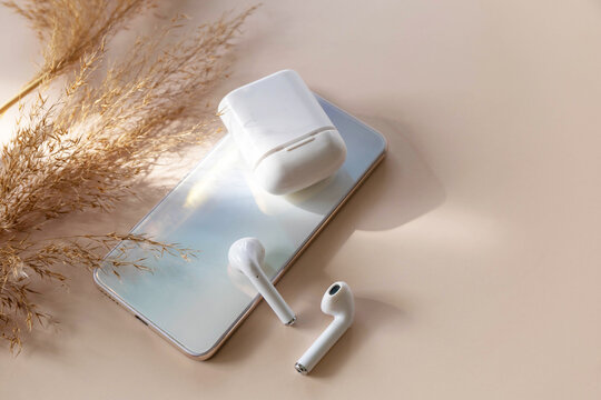 Minimal shot iPhone smartphone and of a pair of white wireless headphones, dried grass on a pastel neutral colors background. Wireless technology, modern tech. Copy space.