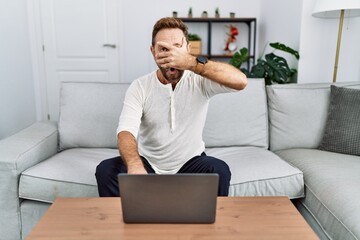 Middle age man using laptop at home peeking in shock covering face and eyes with hand, looking through fingers with embarrassed expression.