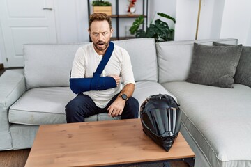 Middle age man with beard wearing arm on sling because motorbike accident scared and amazed with...