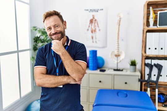 Middle age physiotherapist man working at pain recovery clinic looking confident at the camera smiling with crossed arms and hand raised on chin. thinking positive.