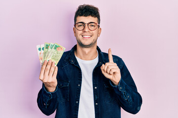 Young hispanic man holding argentine pesos banknotes smiling with an idea or question pointing...