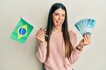 Young hispanic woman holding brazil flag and real banknotes winking looking at the camera with sexy...