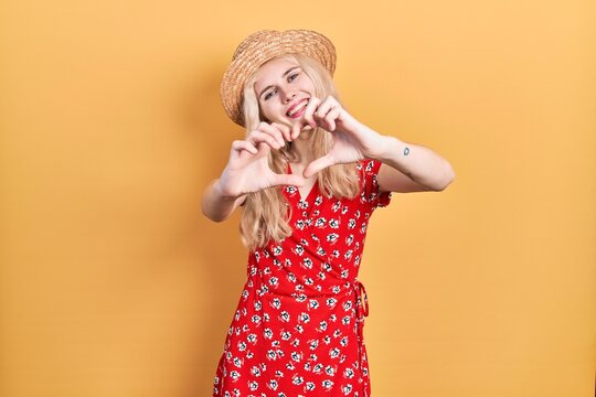 Beautiful caucasian woman with blond hair wearing summer hat smiling in love doing heart symbol shape with hands. romantic concept.