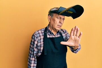 Handsome senior man with grey hair wearing welding protection mask doing stop sing with palm of the...